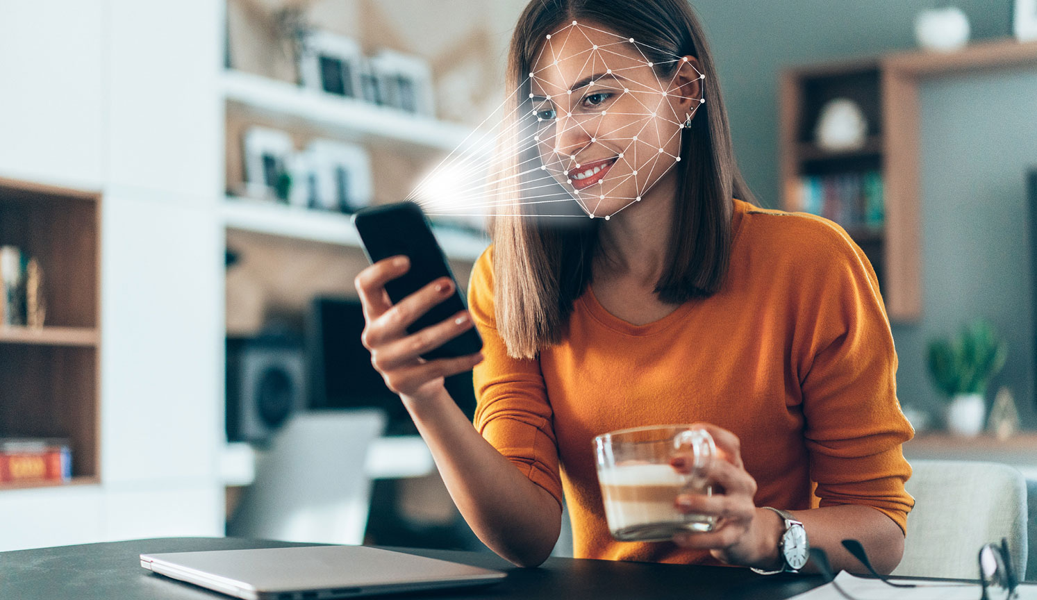 smiling woman in orange sitting at desk, holding smartphone which is digitally scanning her face